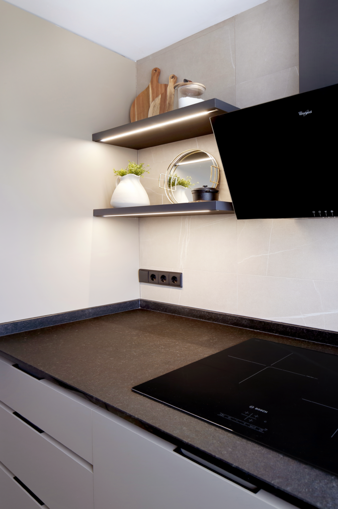 Rincón de cocina con encimera negra, baldas con iluminación LED encendida y objetos decorativos encima, y con la campana extractora y la vitrocerámica de cristal negro. Diseñado en un proyecto de interiorismo