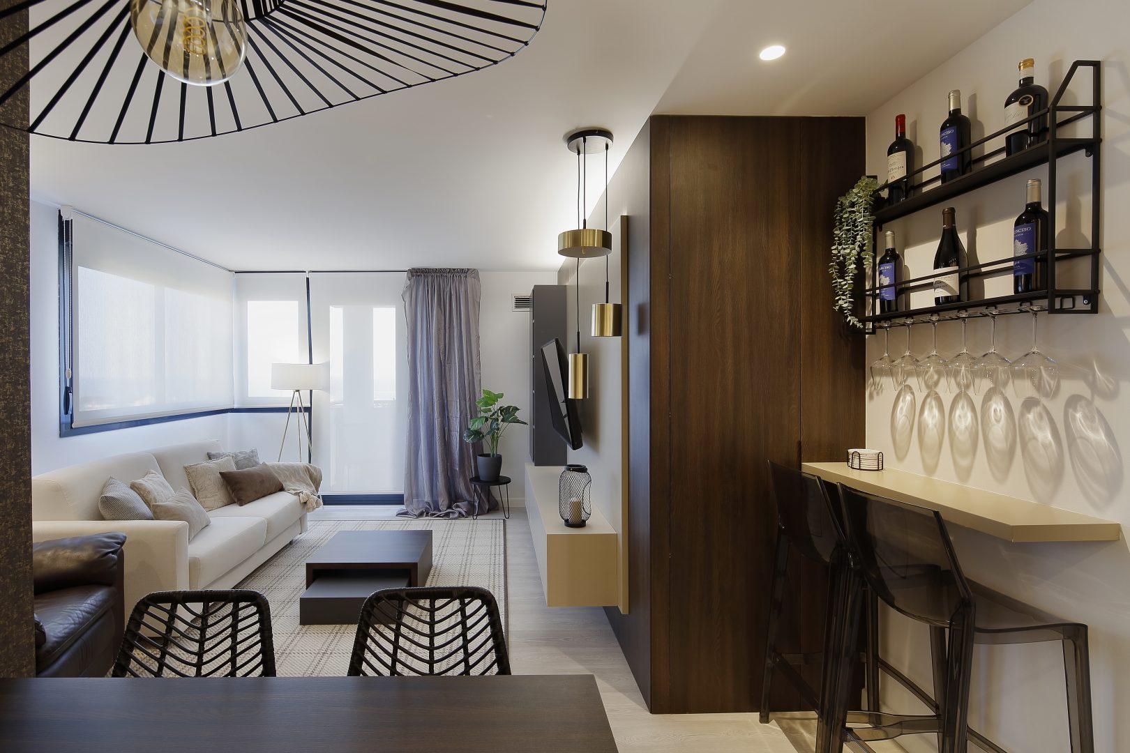 Espacio de salón con zona de barra de bar y botellas en pared. Panel de madera junto a los taburetes, diseñado en proyecto de interiorismo